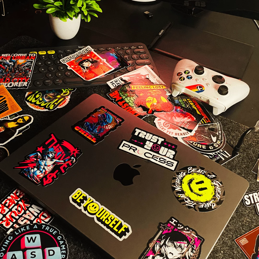 Tampa de notebook cinza-escuro coberta de adesivos recortados no contorno, sobre uma mesa com mais adesivos
