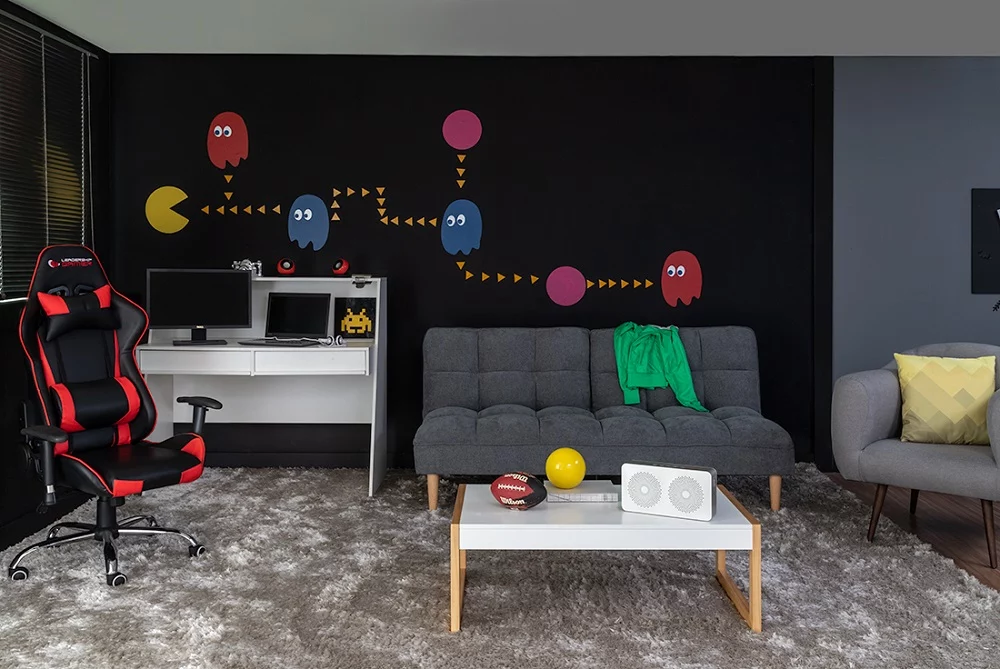 Decoração nerd numa sala de parede preta com Pac-Man e fantasmas, cadeira gamer e sofá cinza