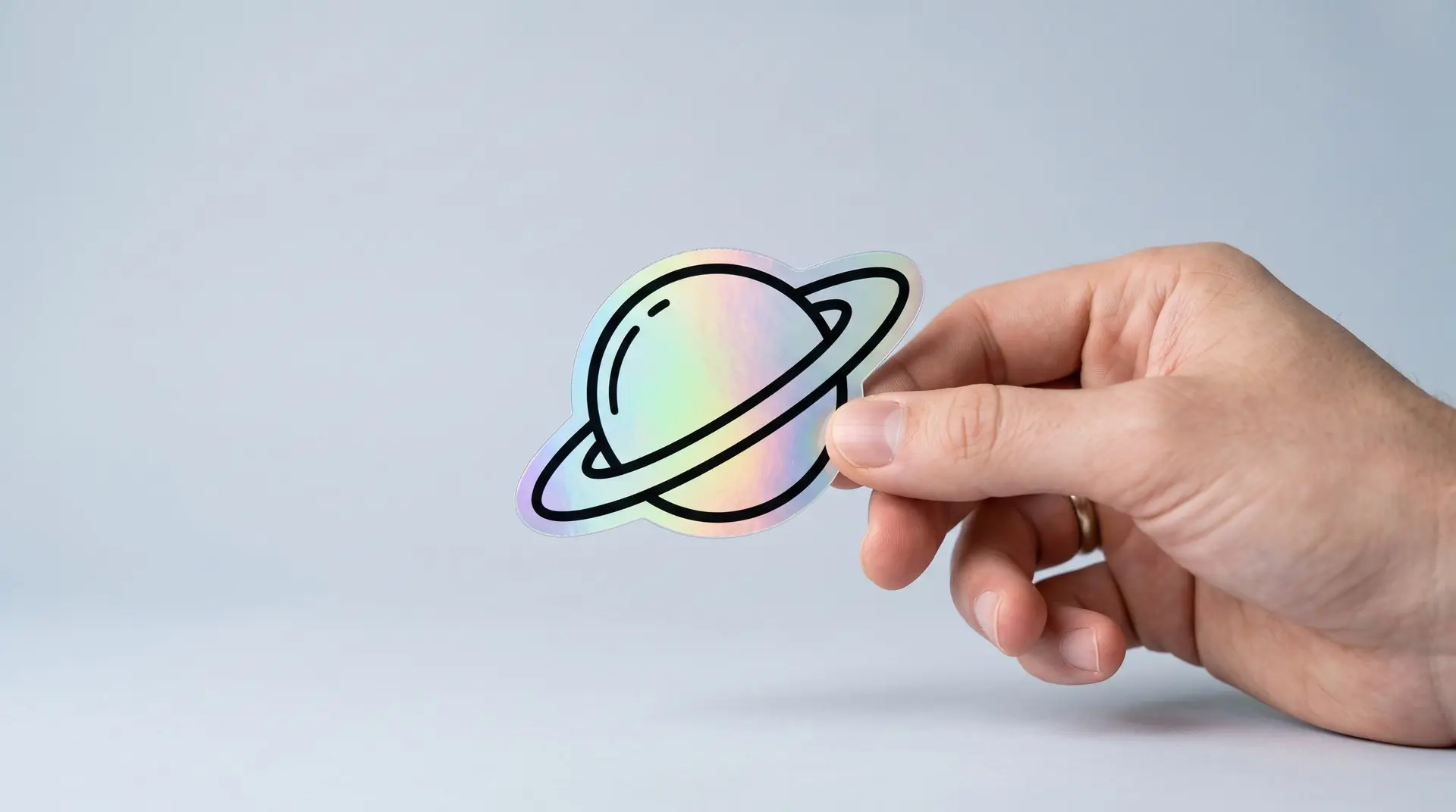 Mão segurando adesivo holográfico de planeta com reflexo de arco-íris em fundo cinza claro